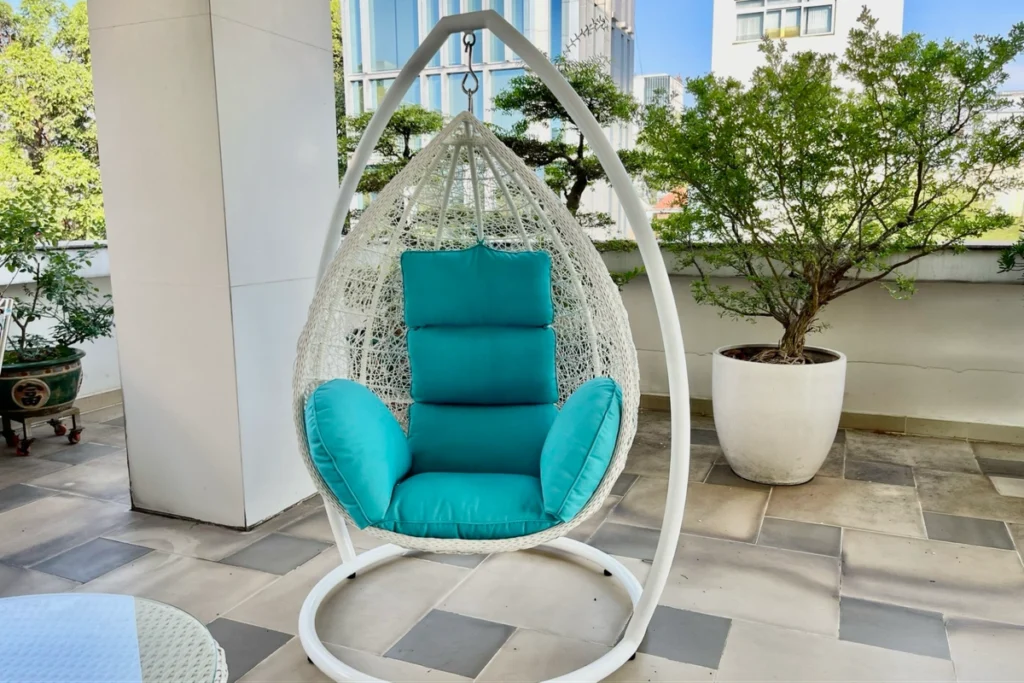 15 Ý Tưởng Decor Ban Công Đẹp Cho Cuộc Sống Thêm Chill 13 Ghế xích đu ngoài trời khung sắt trắng với đệm xanh nổi bật, đặt giữa ban công nhiều cây xanh tạo cảm giác thư giãn.