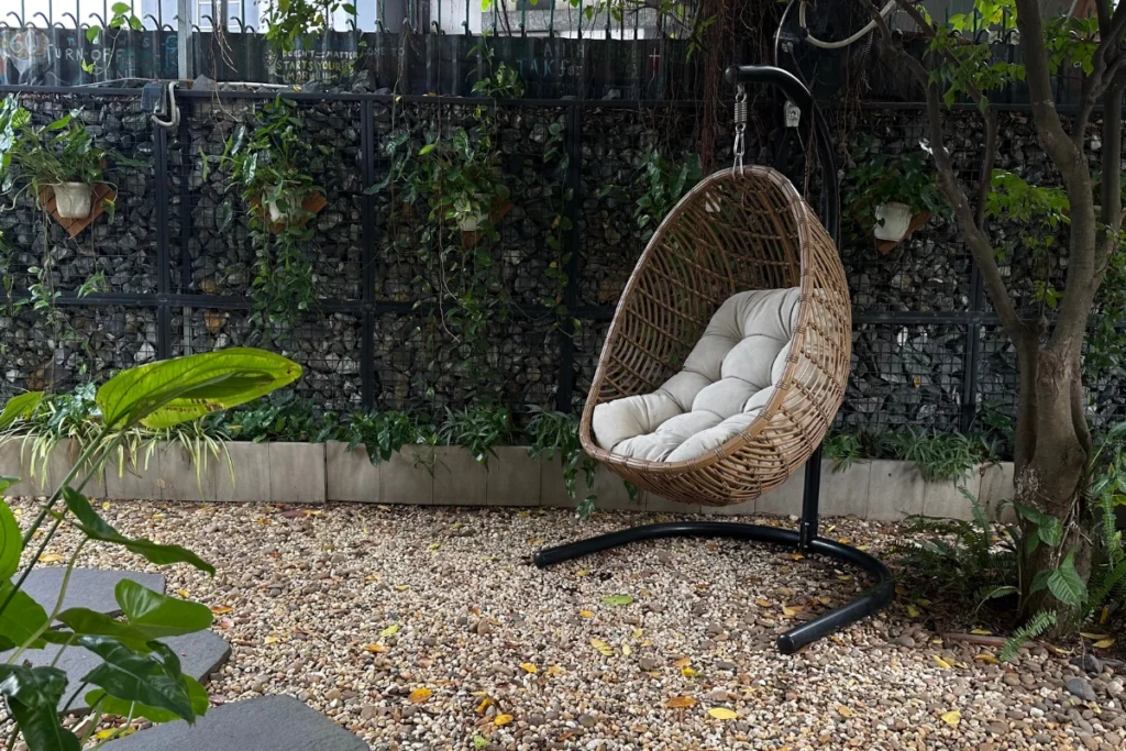 Các cách tạo ra sân vườn đẹp tự làm tại nhà – Biến mọi góc nhỏ thành không gian chill lý tưởng 2 Ghế xích đu mây tre kèm đệm đặt trong góc sân trải sỏi có cây xanh.