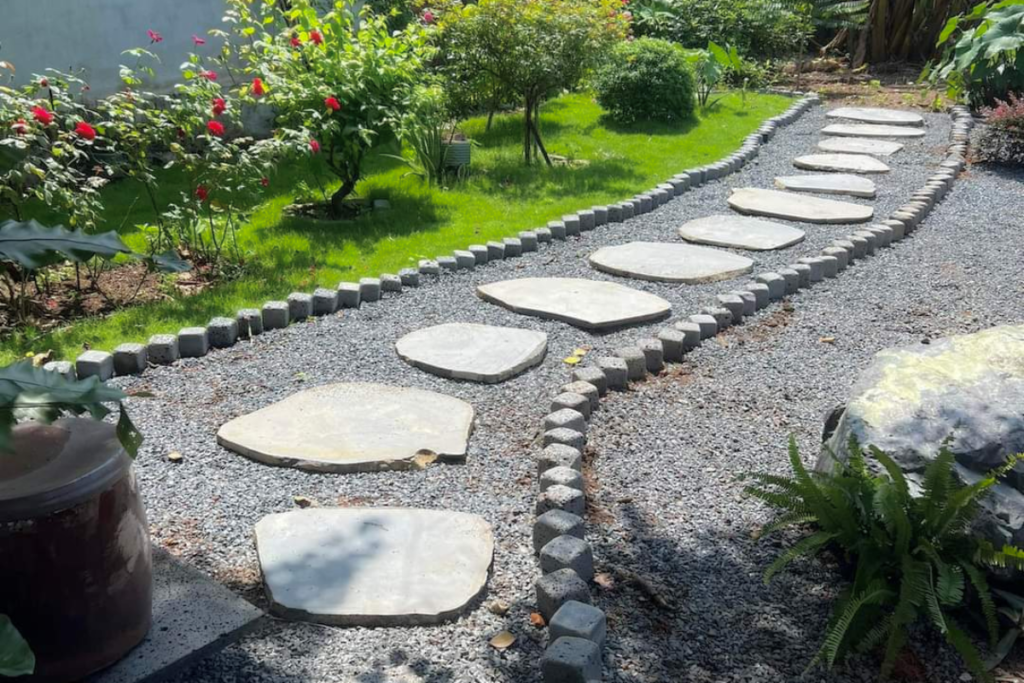 Top 7 loại đá lát nền sân vườn siêu bền đẹp theo thời gian 6 Lối đi sân vườn lát bằng đá hoa cương với các phiến đá tự nhiên sắp xếp xen kẽ trên nền sỏi, bao quanh bởi cây xanh và hoa tươi.