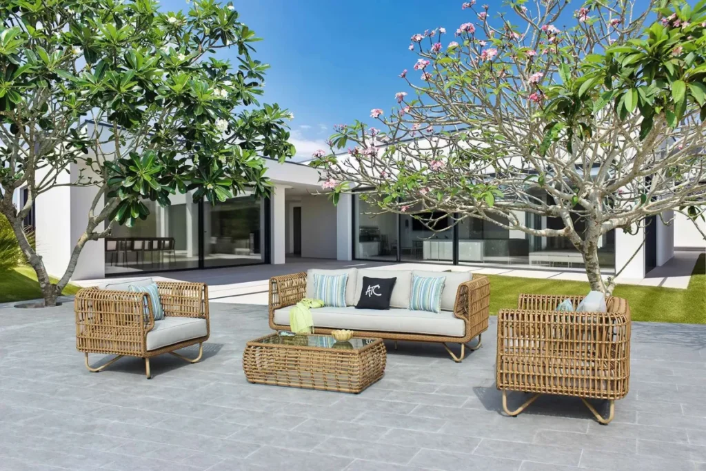 Sân vườn ngoài trời với cây lớn trồng cố định, bộ bàn ghế mây đặt trên nền gạch, tạo bố cục hài hòa và ổn định.