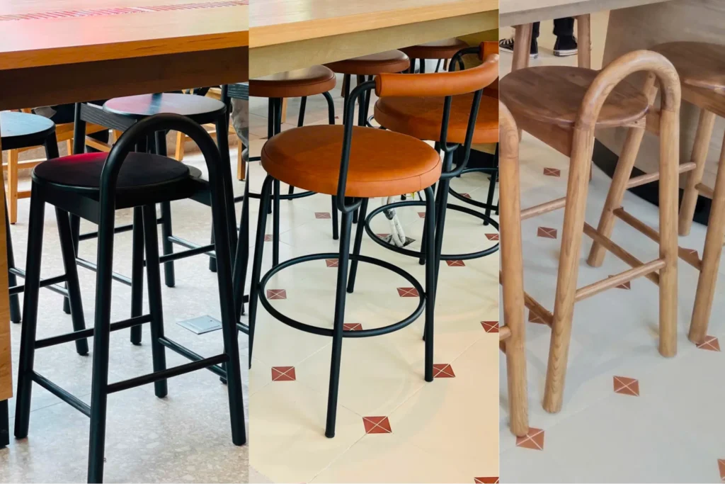 Các mẫu ghế quầy bar với khung kim loại sơn đen và mặt ngồi gỗ, đặt dưới quầy bar trong không gian quán cà phê hiện đại.