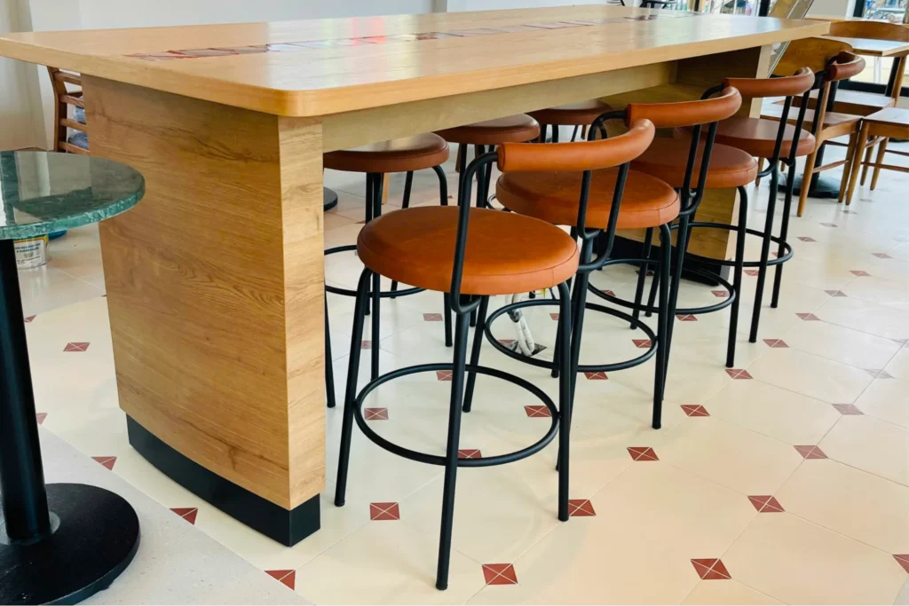 ghế ngồi quầy bar có tựa lưng, khung kim loại sơn đen kết hợp mặt ngồi và tựa lưng gỗ, bố trí dọc theo quầy bar gỗ trong không gian quán.